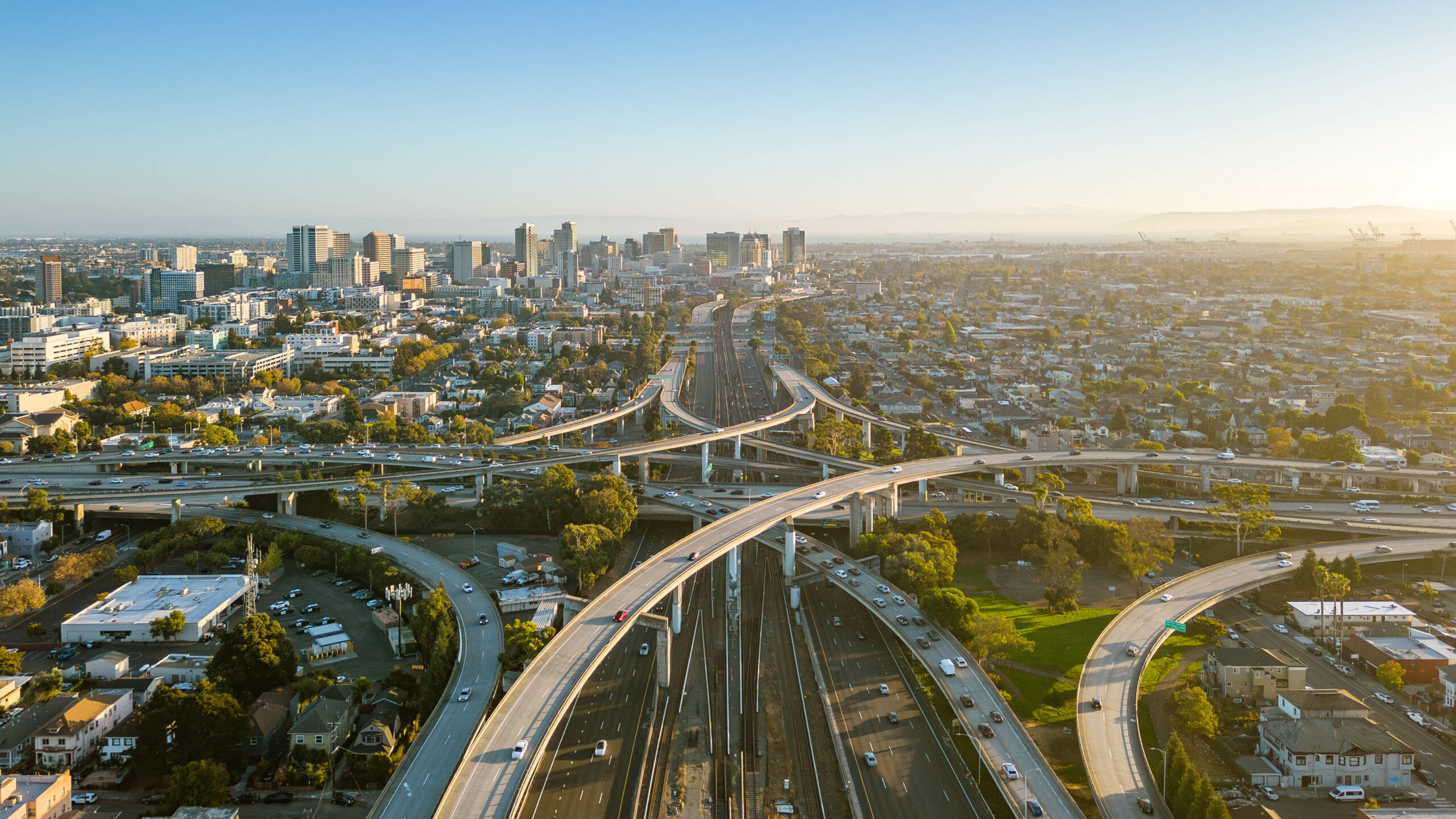Aerial view of a sprawling highway interchange with multiple overpasses and cars, set against a city skyline under a clear blue sky at sunset.