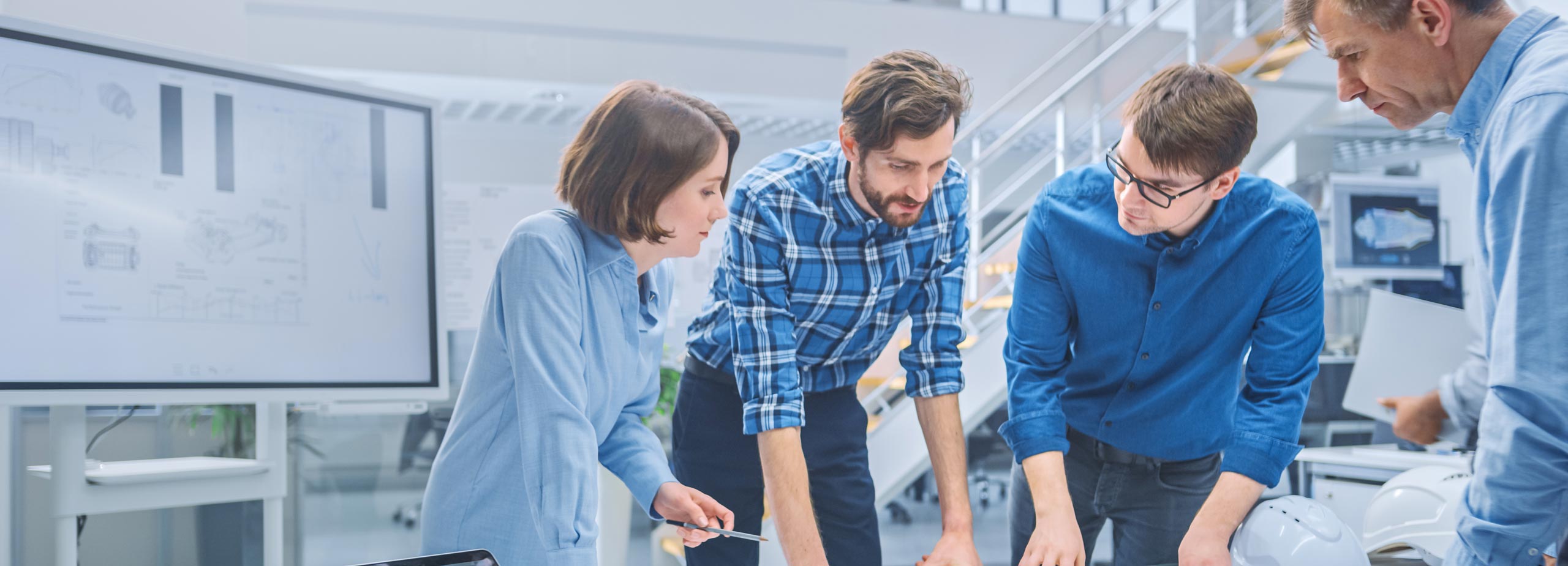 Four people in an office, gathered around a table, discussing documents. They appear focused and engaged. A large monitor with diagrams is visible in the background, and the setting is modern and bright.