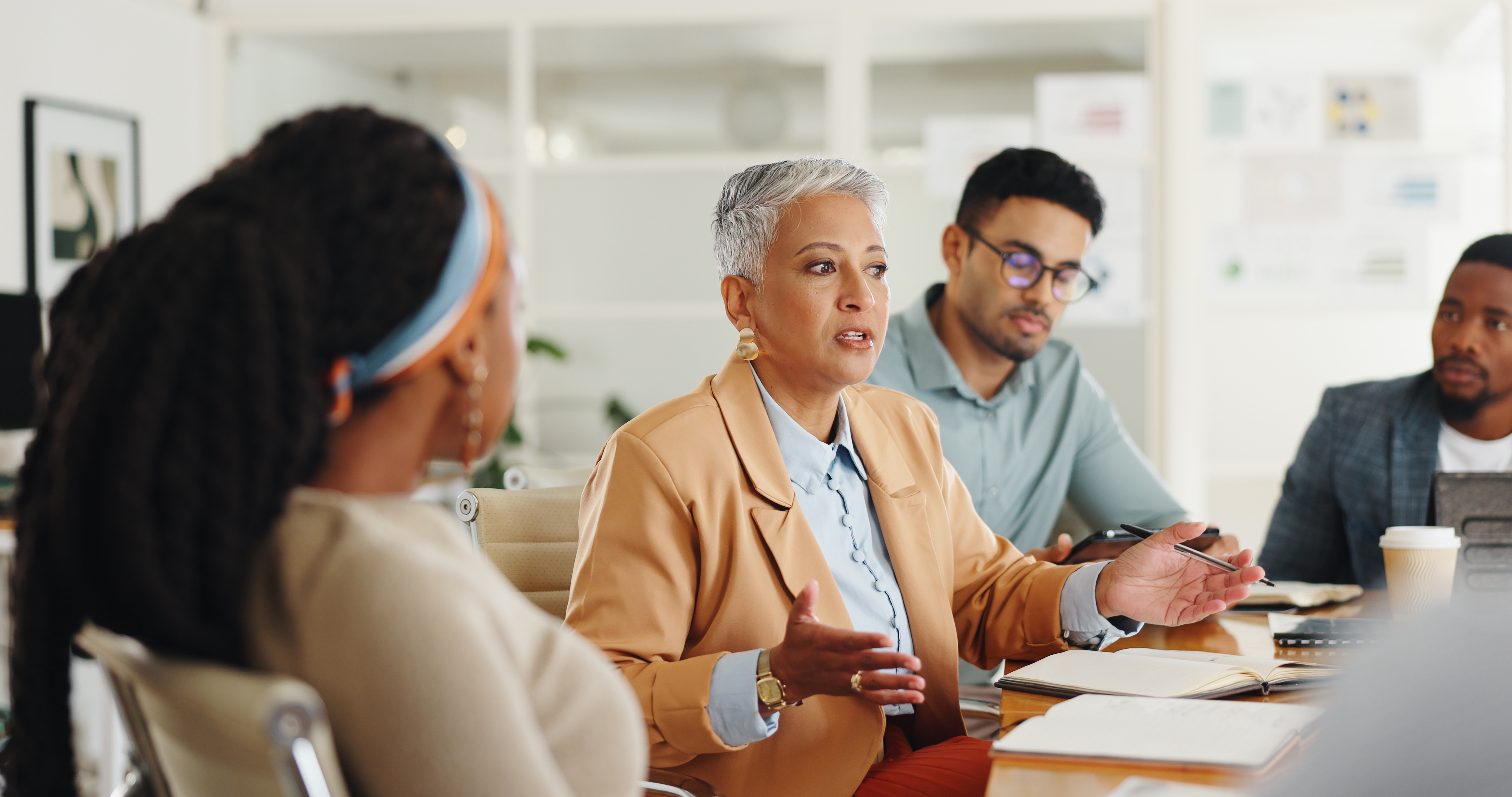 A woman with short gray hair speaks during a meeting at a conference table with three colleagues, who listen attentively. There are notebooks and papers on the table in a bright office setting.