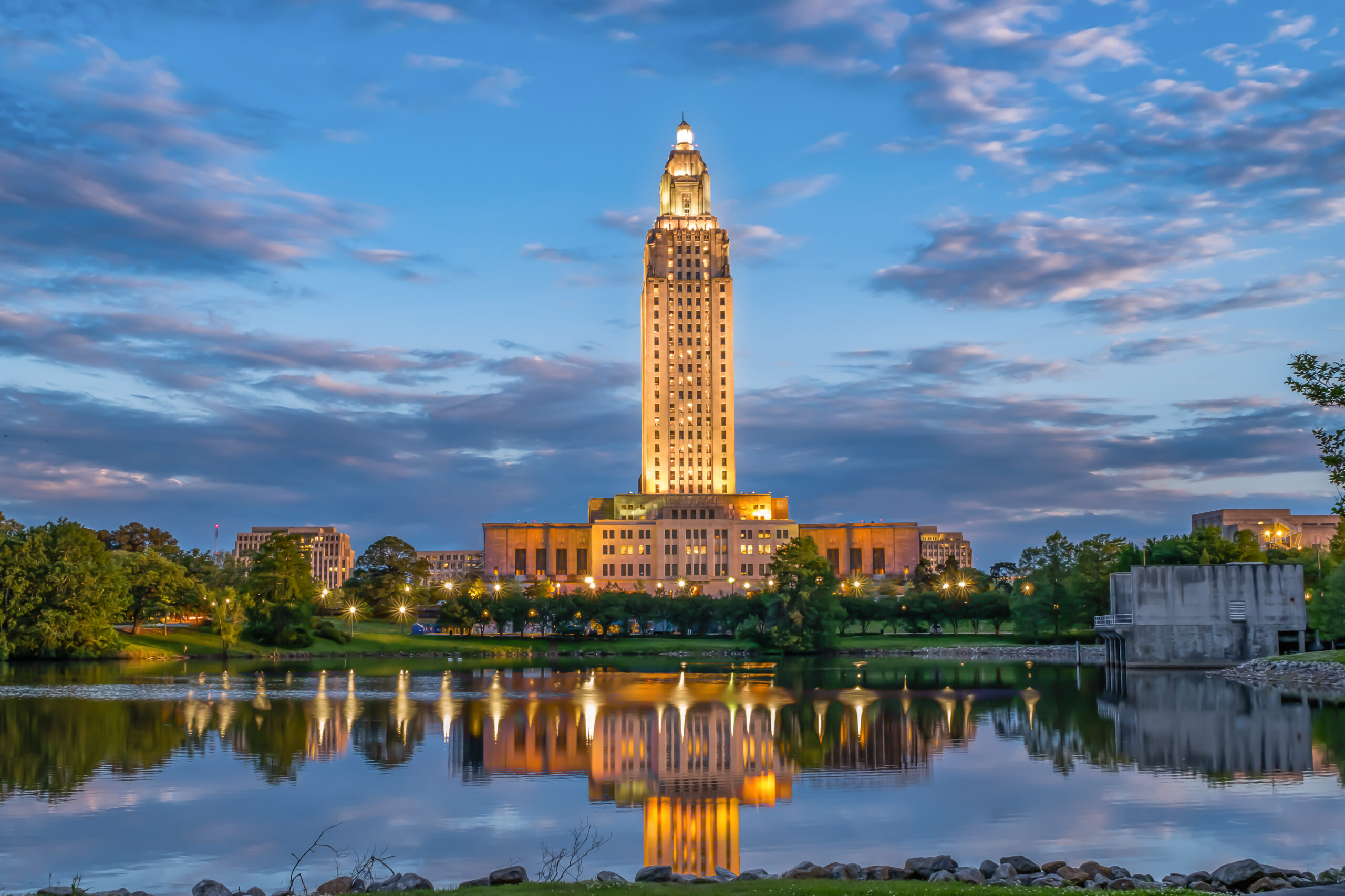 A tall, illuminated art deco building stands against a twilight sky, reflected in a calm lake surrounded by trees and city lights.