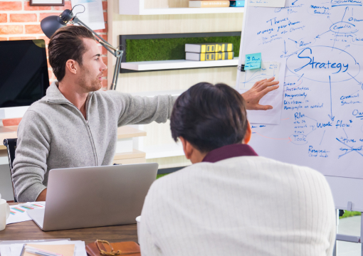 Two people in a modern office discuss a strategy plan. One gestures toward a whiteboard covered in handwritten notes and diagrams, while the other listens. A laptop and office supplies are visible on the table.