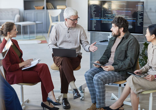 Four people sit in a circle having a discussion in an office. One older man gestures while speaking, holding a laptop. A screen behind them displays computer code. The others listen, holding tablets and notebooks.