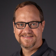 A man with short brown hair, glasses, and a short beard smiles at the camera against a dark background. He is wearing a dark collared shirt.