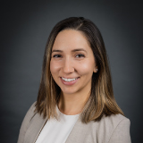 A woman with straight, shoulder-length brown hair smiles at the camera. She is wearing a light beige blazer over a white top, and the background is a dark gray gradient.